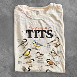 May include: A cream-colored t-shirt featuring the text Stop staring at my TITS in black and orange. The design includes detailed illustrations of various tit bird species, including coal, blue, willow, great, crested, marsh, and varied tits.