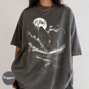 Puede incluir: Una camiseta gris carbón de manga corta con un estampado gráfico de una criatura alada con ojos rojos brillantes, una luna llena, nubes y un paisaje urbano. El texto "POINT PLEASANT WEST VIRGINIA" está impreso en la esquina inferior derecha.