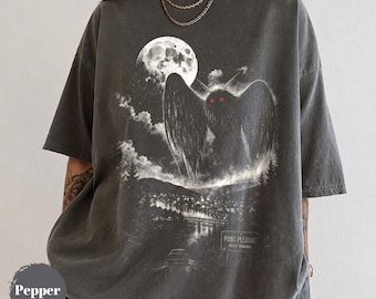 Könnte beinhalten: Ein anthrazitfarbenes Kurzarm-T-Shirt mit einem Grafikdruck einer geflügelten Kreatur mit leuchtend roten Augen, einem Vollmond, Wolken und einer Stadtlandschaft. Der Text "POINT PLEASANT WEST VIRGINIA" ist unten rechts auf der Grafik aufgedruckt.