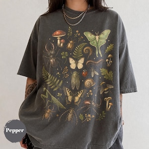 以下が含まれることがあります： チャコールグレーの半袖Tシャツで、様々な昆虫、蝶、キノコ、シダが描かれた詳細なグラフィックプリントが施されています。デザインには、大きな緑色の蛾、クワガタ、カタツムリ、カマキリが含まれています。Pepperという文字が見えます。