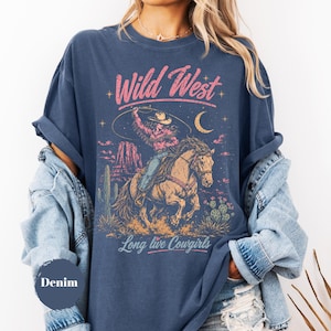 May include: A denim blue short-sleeve t-shirt featuring a graphic print of a cowgirl riding a horse with a lasso. The design includes desert scenery, cacti, a crescent moon, and the text Wild West and Long live Cowgirls in pink and white.