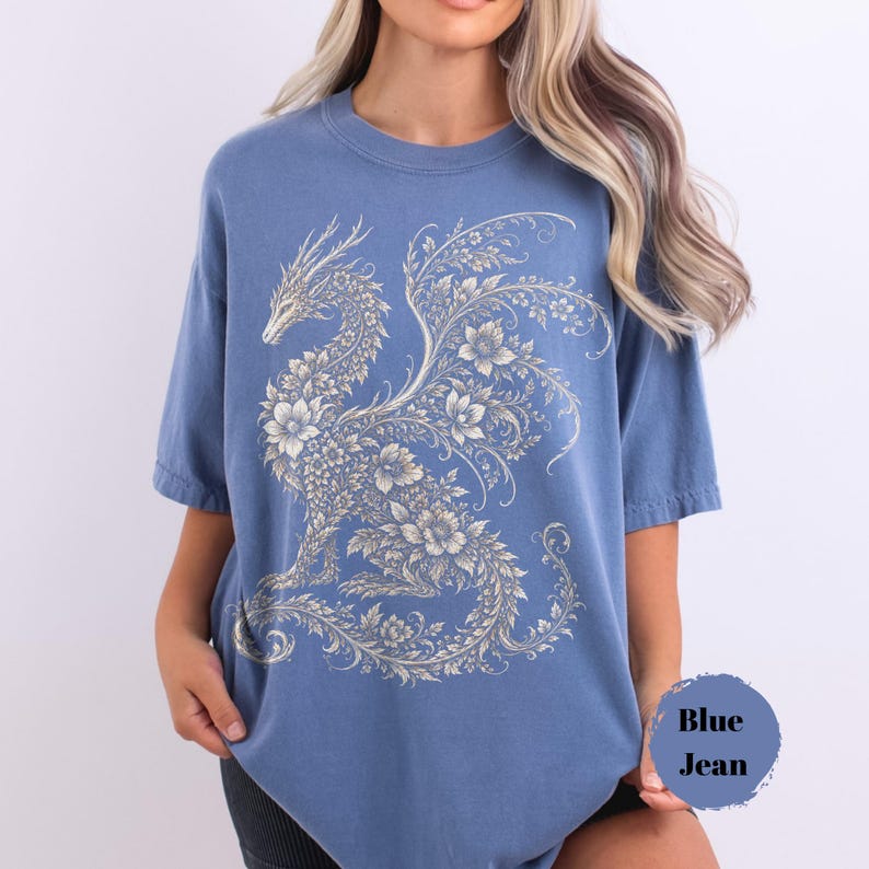 May include: A blue jean colored t-shirt featuring a large, intricate graphic print of a dragon composed entirely of floral and vine patterns in a light cream color. The shirt has a crew neckline, short sleeves, and a relaxed, loose-fitting silhouette.
