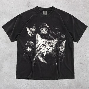 K&ouml;nnte beinhalten: Ein schwarzes kurz&auml;rmeliges T-Shirt mit einem wei&szlig;en Grafikdruck von mehreren Katzen, von denen einige Kapuzenpullover tragen, wobei eine zentrale Katze eine Zigarette im Mund zu haben scheint. Das Shirt hat einen Rundhalsausschnitt und ein verwaschenes Finish.