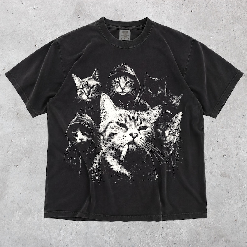 K&ouml;nnte beinhalten: Ein schwarzes kurz&auml;rmeliges T-Shirt mit einem wei&szlig;en Grafikdruck von mehreren Katzen, von denen einige Kapuzenpullover tragen, wobei eine zentrale Katze eine Zigarette im Mund zu haben scheint. Das Shirt hat einen Rundhalsausschnitt und ein verwaschenes Finish.
