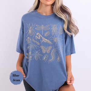 May include: A blue t-shirt featuring a gold-colored graphic print of various insects, including a beetle, dragonfly, praying mantis, spider, butterfly, bee, and centipede, along with botanical elements like mushrooms and leaves.