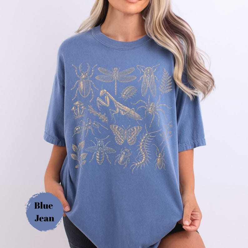 May include: A blue t-shirt featuring a gold-colored graphic print of various insects, including a beetle, dragonfly, praying mantis, spider, butterfly, bee, and centipede, along with botanical elements like mushrooms and leaves.