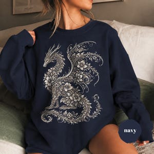 May include: A navy blue crewneck sweatshirt featuring a large, intricate graphic of a dragon composed entirely of detailed floral patterns and swirling vines in shades of white and light gray. The design covers the center of the chest area.