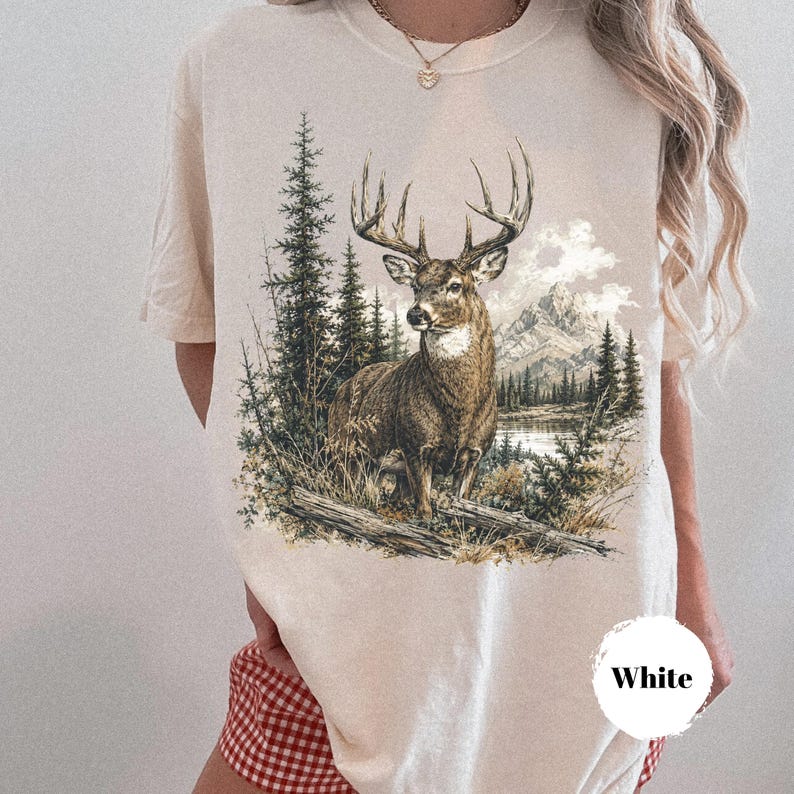 K&ouml;nnte beinhalten: Ein cremefarbenes T-Shirt mit einem detaillierten Grafikdruck eines gro&szlig;en Hirsches mit Geweih, der in einer Waldlandschaft mit Tannen, einem See und Bergen im Hintergrund steht. Das T-Shirt hat einen Rundhalsausschnitt und kurze &Auml;rmel.