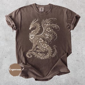 May include: A brown short-sleeve t-shirt featuring a large, intricate graphic of a dragon composed entirely of floral and vine patterns in a light beige color. The shirt has a crew neckline and a small label at the inner collar. The word Espresso is visible.