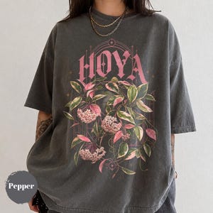 May include: A charcoal gray short-sleeve t-shirt featuring a graphic print of a Hoya plant with pink and green variegated leaves and clusters of small pink flowers. The word HOYA is printed in a large, stylized pink font above the botanical illustration.