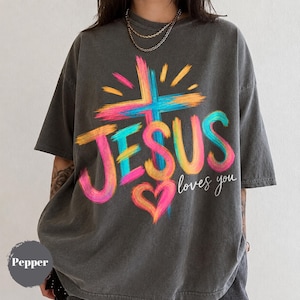 Puede incluir: Una camiseta gris carbón de manga corta con un gráfico colorido de una cruz, un corazón y el texto "JESUS loves you" en letras estilo pincelada. El diseño utiliza tonos vibrantes de rosa, azul y amarillo sobre la tela oscura.