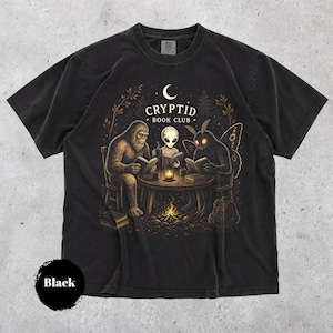 May include: A black short-sleeved t-shirt featuring a graphic print of Bigfoot, an alien, and a mothman sitting at a round table reading books. The text "CRYPTID BOOK CLUB" is printed above the figures, with a crescent moon and a campfire below.