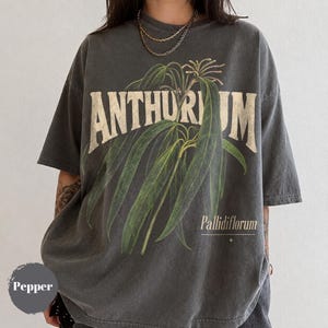 May include: A charcoal gray short-sleeve t-shirt featuring a large botanical illustration of an Anthurium plant with long green leaves. The text ANTHURIUM is printed in large cream letters above the plant, with Pallidiflorum printed below.
