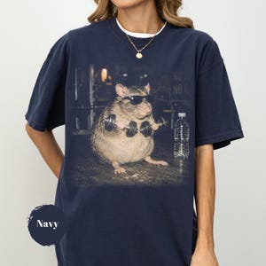 May include: A navy blue crewneck t-shirt featuring a graphic print of a rat wearing black sunglasses and lifting small dumbbells in a gym setting, with a clear plastic water bottle placed next to the rat on the floor.