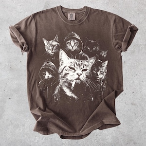 K&ouml;nnte beinhalten: Ein braunes Kurzarm-T-Shirt mit einem wei&szlig;en Grafikdruck von mehreren Katzen, von denen einige Kapuzenpullover tragen. Die zentrale Katze ist mit einer Zigarette im Mund dargestellt. Das Shirt hat einen Rundhalsausschnitt und umgekrempelte &Auml;rmel.