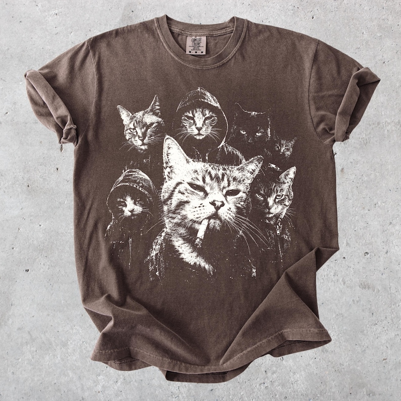 K&ouml;nnte beinhalten: Ein braunes Kurzarm-T-Shirt mit einem wei&szlig;en Grafikdruck von mehreren Katzen, von denen einige Kapuzenpullover tragen. Die zentrale Katze ist mit einer Zigarette im Mund dargestellt. Das Shirt hat einen Rundhalsausschnitt und umgekrempelte &Auml;rmel.
