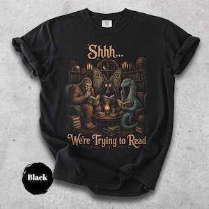 Könnte beinhalten: Ein schwarzes T-Shirt mit einer grafischen Illustration von Bigfoot, einem mottenartigen Wesen und einem langhalsigen Dinosaurier, die in einer Bibliothek bei Laternenlicht lesen. Der Text oben lautet Shhh... und der Text unten We're Trying to Read.