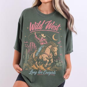 May include: An olive green t-shirt featuring a graphic print of a cowgirl riding a horse while swinging a lasso. The design includes the text "Wild West" in pink and "Long live Cowgirls" in light blue, with desert scenery and a crescent moon.