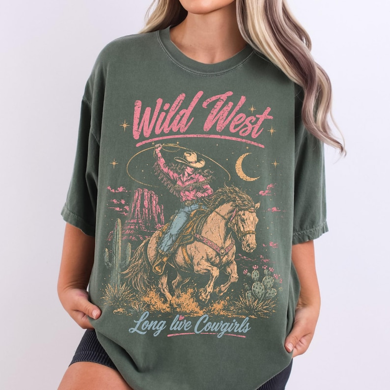 May include: An olive green t-shirt featuring a graphic print of a cowgirl riding a horse while swinging a lasso. The design includes the text "Wild West" in pink and "Long live Cowgirls" in light blue, with desert scenery and a crescent moon.