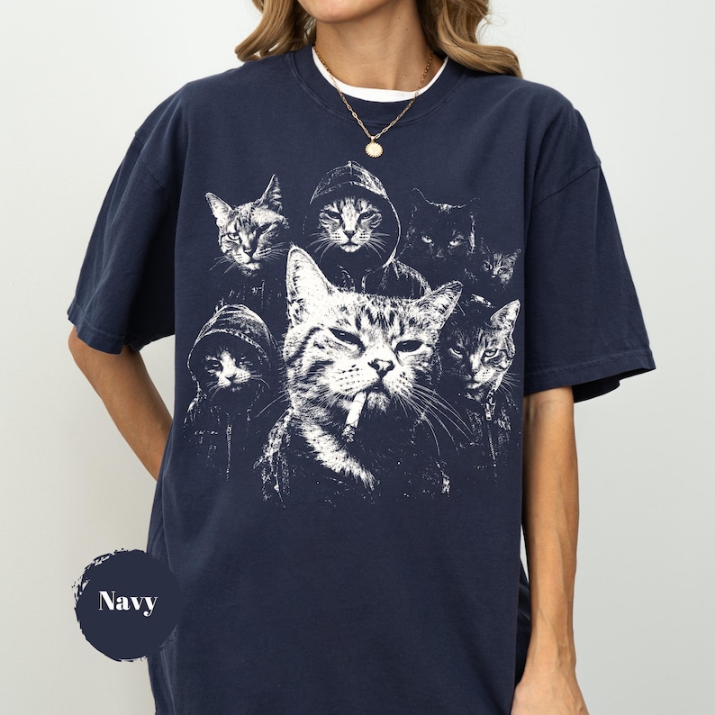 K&ouml;nnte beinhalten: Ein kurz&auml;rmeliges T-Shirt in Marineblau mit einem wei&szlig;en Grafikdruck von mehreren Katzen, von denen einige Kapuzenpullover tragen, wobei eine zentrale Katze eine Zigarette zu rauchen scheint. Das T-Shirt hat einen Rundhalsausschnitt und eine lockere Passform.