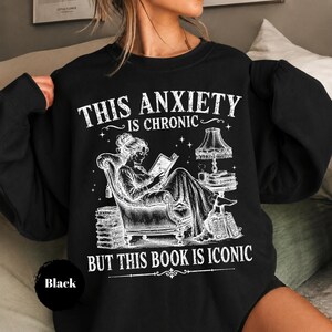 May include: A black crewneck sweatshirt featuring a white graphic illustration of a person reading in an armchair next to a lamp and stacks of books. The text reads: THIS ANXIETY IS CHRONIC BUT THIS BOOK IS ICONIC. The design has a distressed texture.