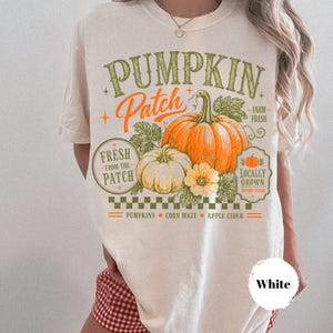 Może przedstawiać: Kremowy t-shirt z nadrukiem graficznym z tekstem PUMPKIN PATCH, FARM FRESH, FRESH FROM THE PATCH, LOCALLY GROWN, PUMPKINS, CORN MAZE oraz APPLE CIDER. Wzór zawiera ilustracje pomarańczowych i białych dyń oraz liści.
