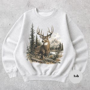 K&ouml;nnte beinhalten: Ein hellgraues Sweatshirt mit Rundhalsausschnitt und einem detaillierten Grafikdruck eines braunen Hirsches mit gro&szlig;em Geweih, der in einer Waldlandschaft mit Tannen und Bergen im Hintergrund steht. Das Wort Ash steht in einem kleinen Kreis.