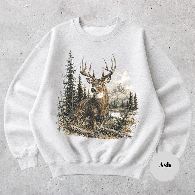 K&ouml;nnte beinhalten: Ein hellgraues Sweatshirt mit Rundhalsausschnitt und einem detaillierten Grafikdruck eines braunen Hirsches mit gro&szlig;em Geweih, der in einer Waldlandschaft mit Tannen und Bergen im Hintergrund steht. Das Wort Ash steht in einem kleinen Kreis.