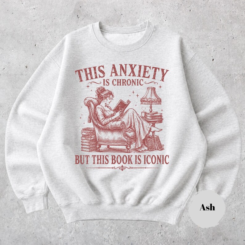 May include: An ash gray crewneck sweatshirt featuring a red graphic illustration of a person reading in an armchair next to a lamp and stacks of books. The text reads: THIS ANXIETY IS CHRONIC BUT THIS BOOK IS ICONIC. The design is centered on the chest.