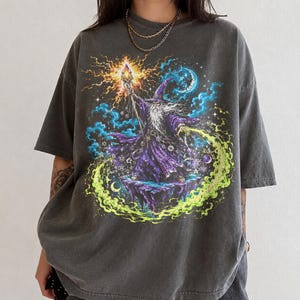 May include: A charcoal gray short-sleeve t-shirt featuring a graphic print of a wizard in a purple robe holding a glowing staff. The design includes a crescent moon, swirling blue and green energy, and celestial elements against a dark background.
