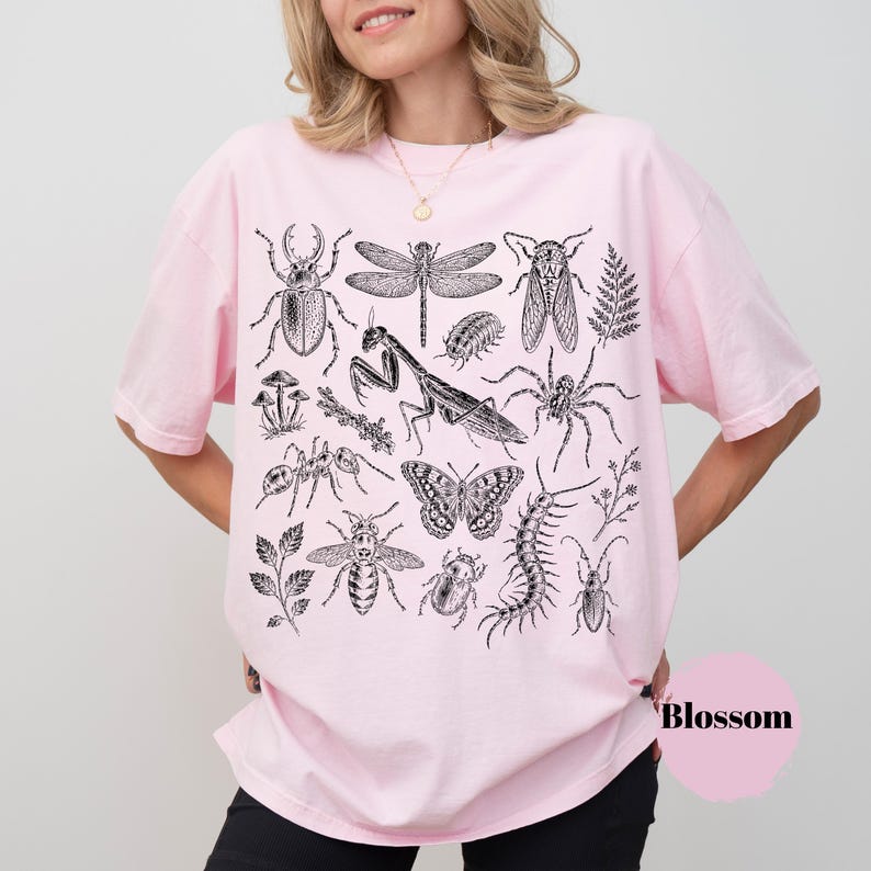 May include: A light pink short-sleeve t-shirt featuring a detailed black ink-style graphic print of various insects, including a praying mantis, dragonfly, butterfly, beetle, and spider, along with small botanical elements like ferns and mushrooms.