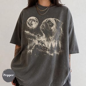Könnte beinhalten: Ein anthrazitfarbenes Kurzarm-T-Shirt mit einem Grafikdruck eines Mopses, der auf einer felsigen Klippe steht, vor einem großen Vollmond und Tannenbäumen. Das Design zeigt zusätzlich ein großes, detailliertes Porträt eines Mopsgesichts.