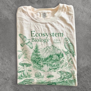 Pode incluir: Uma t-shirt de cor creme com uma impressão gráfica verde com o texto Ecosystem Biology Life in Balance. O design inclui ilustrações de um falcão, uma coruja, uma paisagem montanhosa, cogumelos, um caracol, uma abelha, um sapo e células.