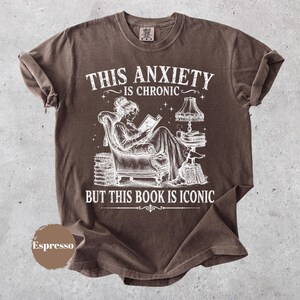 May include: A brown short-sleeve t-shirt featuring a white graphic of a person reading in an armchair with a lamp and stacks of books. The text reads: THIS ANXIETY IS CHRONIC BUT THIS BOOK IS ICONIC. A circular brown swatch labeled Espresso is included.