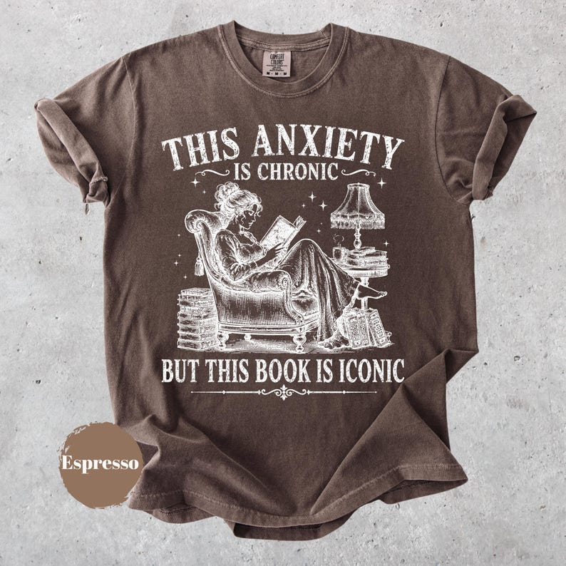 May include: A brown short-sleeve t-shirt featuring a white graphic of a person reading in an armchair with a lamp and stacks of books. The text reads: THIS ANXIETY IS CHRONIC BUT THIS BOOK IS ICONIC. A circular brown swatch labeled Espresso is included.