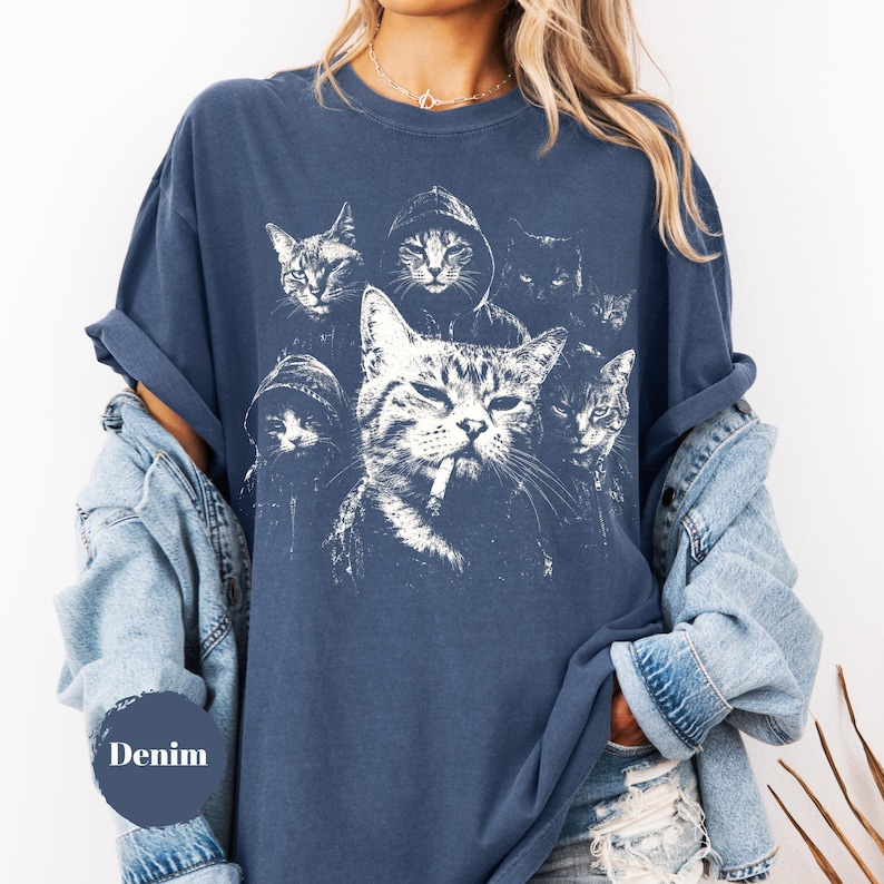 K&ouml;nnte beinhalten: Ein denimfarbenes Kurzarm-T-Shirt mit einem wei&szlig;en Grafikdruck von mehreren Katzen, darunter einige mit Kapuzenpullovern und eine mit einer Zigarette im Mund. Das T-Shirt hat einen Rundhalsausschnitt und eine lockere Passform, getragen unter einer hellblauen Jeansjacke.