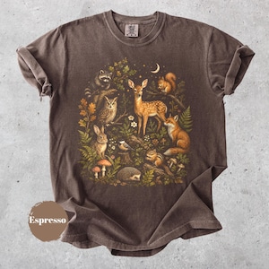 以下が含まれることがあります： ダークブラウンの半袖Tシャツ。鹿、狐、アライグマ、フクロウ、リス、ウサギ、ハリネズミ、シマリスなどの森の動物たちが、キノコ、シダ、オークの葉、三日月に囲まれて描かれた詳細なグラフィックプリントが施されています。