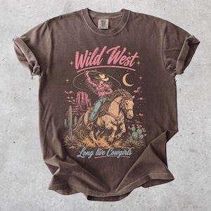 May include: A brown short-sleeve t-shirt featuring a graphic print of a cowgirl riding a horse with a lasso. The design includes desert scenery, cacti, a crescent moon, and the text Wild West and Long live Cowgirls in pink and light blue.
