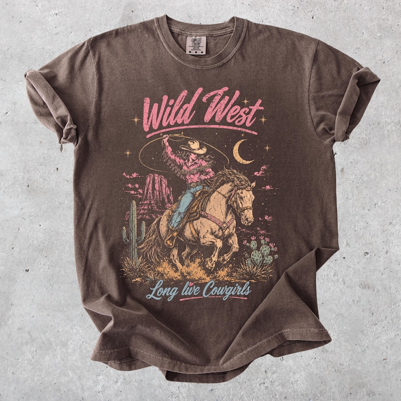 May include: A brown short-sleeve t-shirt featuring a graphic print of a cowgirl riding a horse with a lasso. The design includes desert scenery, cacti, a crescent moon, and the text Wild West and Long live Cowgirls in pink and light blue.