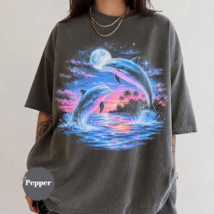 Könnte beinhalten: Ein anthrazitfarbenes Kurzarm-T-Shirt mit einem Grafikdruck von zwei Delfinen, die bei Sonnenuntergang aus dem Ozean springen. Die Szene zeigt einen Vollmond, Palmen und einen lebendigen Himmel in Rosa-, Lila- und Blautönen. Das Wort Pepper ist gedruckt.