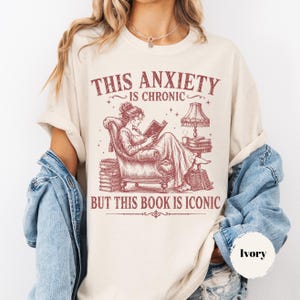 May include: An ivory t-shirt featuring a maroon graphic of a person reading in an armchair with stacks of books and a lamp. The text reads: THIS ANXIETY IS CHRONIC BUT THIS BOOK IS ICONIC. The shirt is layered under a blue denim jacket.