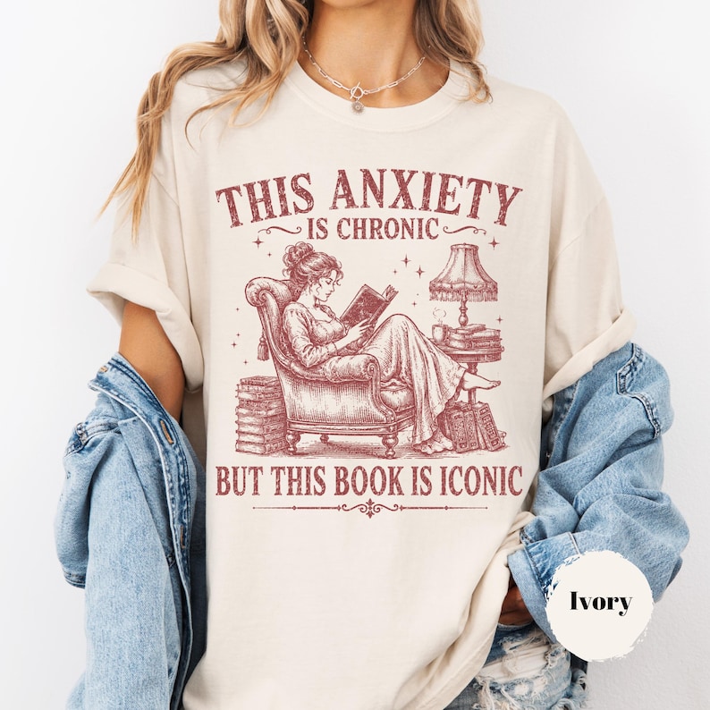 May include: An ivory t-shirt featuring a maroon graphic of a person reading in an armchair with stacks of books and a lamp. The text reads: THIS ANXIETY IS CHRONIC BUT THIS BOOK IS ICONIC. The shirt is layered under a blue denim jacket.