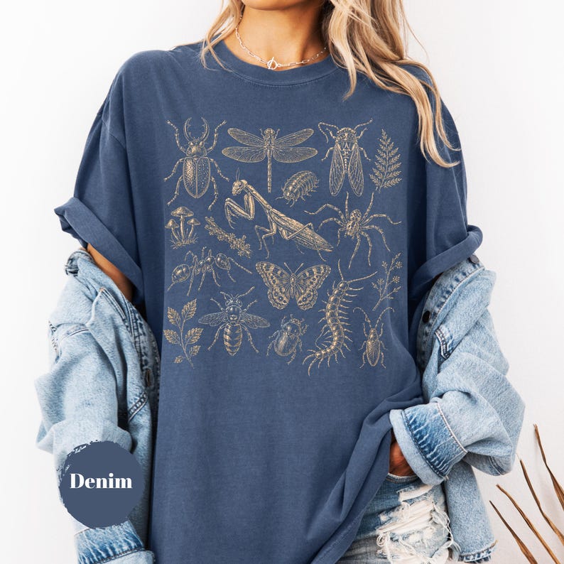 May include: A denim blue t-shirt featuring a gold-toned graphic print of various insects, including a praying mantis, butterfly, dragonfly, beetle, and spider, alongside botanical elements like ferns and mushrooms, with the text Denim.
