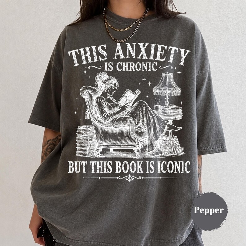 May include: A charcoal gray short-sleeve t-shirt featuring a white graphic of a person reading in an armchair with a lamp and stacks of books. The text reads: THIS ANXIETY IS CHRONIC BUT THIS BOOK IS ICONIC. A small circular label says Pepper.