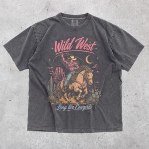 May include: A charcoal grey short-sleeve t-shirt featuring a graphic print of a cowgirl riding a horse in a desert landscape. The text "Wild West" appears in pink above the illustration, and "Long live Cowgirls" is printed in blue below.