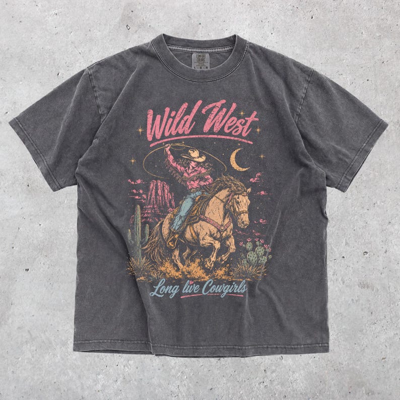 May include: A charcoal grey short-sleeve t-shirt featuring a graphic print of a cowgirl riding a horse in a desert landscape. The text "Wild West" appears in pink above the illustration, and "Long live Cowgirls" is printed in blue below.
