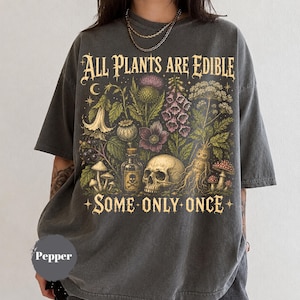 May include: A charcoal gray t-shirt featuring a graphic print with the text "ALL PLANTS ARE EDIBLE SOME ONLY ONCE". The design includes botanical illustrations of flowers, mushrooms, a skull, and a small glass bottle with a skull and crossbones label.
