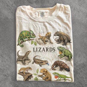 Könnte beinhalten: Ein cremefarbenes T-Shirt mit einem Grafikdruck verschiedener Echsenarten, darunter ein Chamäleon, eine Bartagame und ein Komodowaran. Der Text "LIZARDS" und "ILLUSTRATED SPECIES OF THE NATURAL WORLD" ist in der Mitte des Shirts aufgedruckt.
