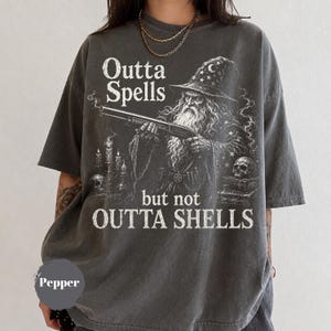 May include: A charcoal grey short-sleeve t-shirt featuring a graphic of a wizard holding a shotgun. The text reads Outta Spells but not OUTTA SHELLS. The design includes a wizard hat, candles, a skull, and books in a white, distressed print.