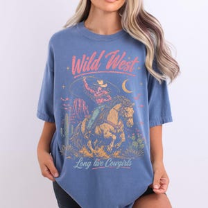 May include: A blue short-sleeve t-shirt featuring a graphic print of a person riding a horse with a lasso. The text "Wild West" appears at the top and "Long live Cowgirls" at the bottom, accompanied by desert imagery including cacti and mesas.